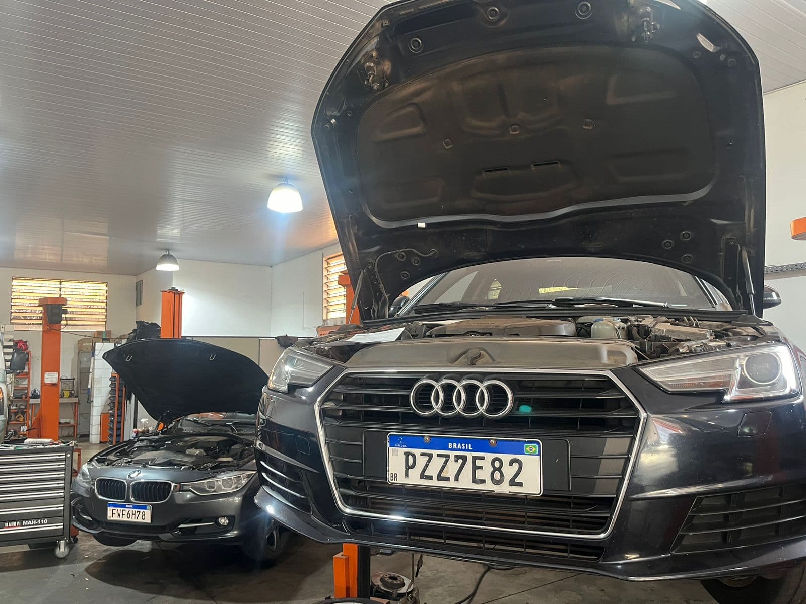 Dentro da Edicar: um Audi e um BMW de capô aberto entre os elevadores laranja da oficina